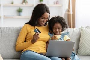 Happy Mother And Little Daughter Shopping Online With Laptop Computer And Credit Card While Relaxing On Couch