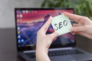 person holding sticky note labeled SEO in front of laptop